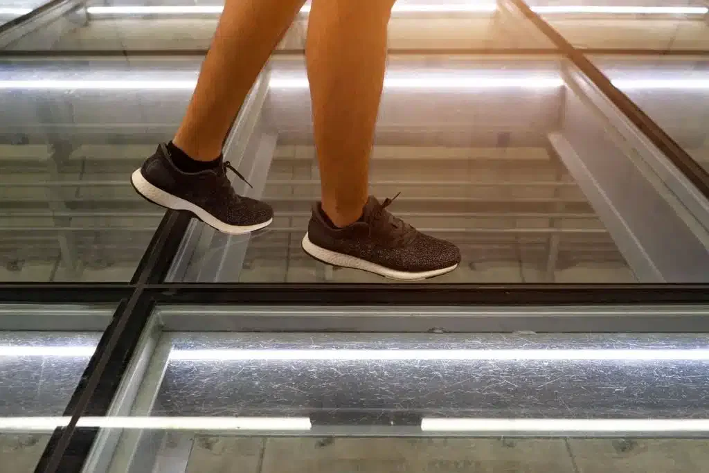Close-up of a person wearing black sneakers walking across a modern, reinforced glass floor with visible support beams and glowing light strips.
