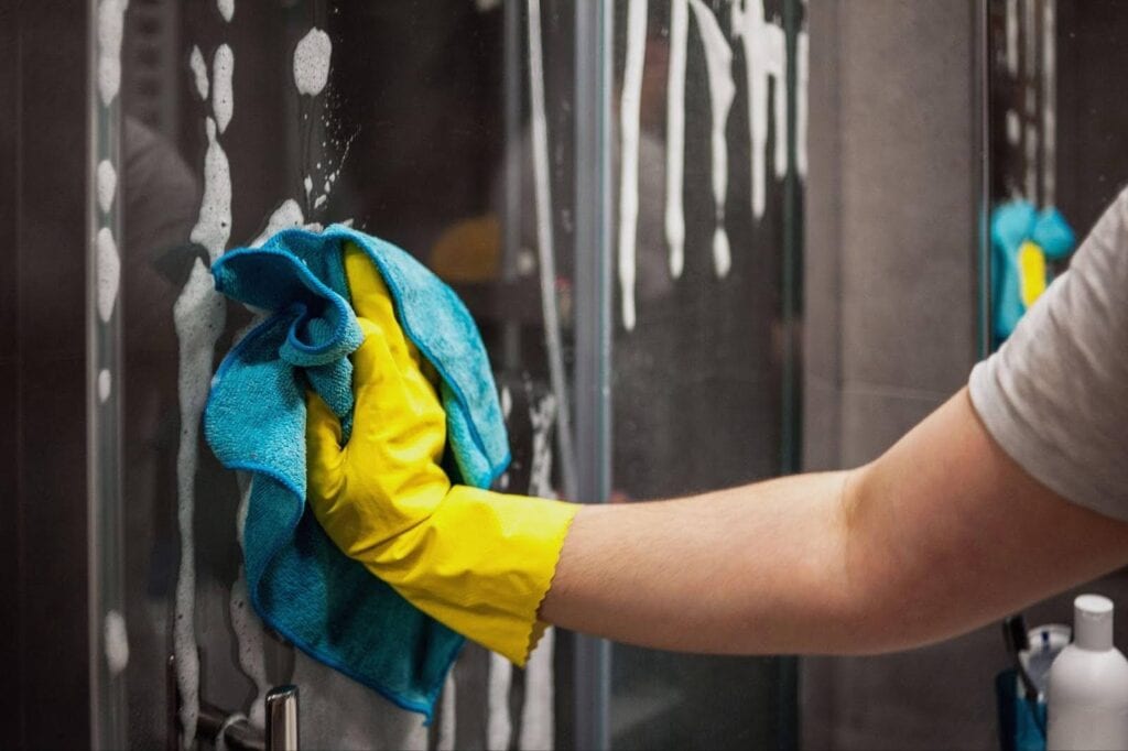 A person in a yellow glove cleans a glass shower door for a streak-free shine