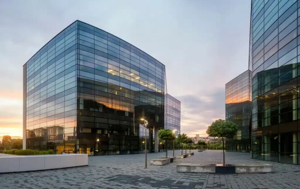 Modern glass office buildings at sunset with a paved courtyard and small trees.