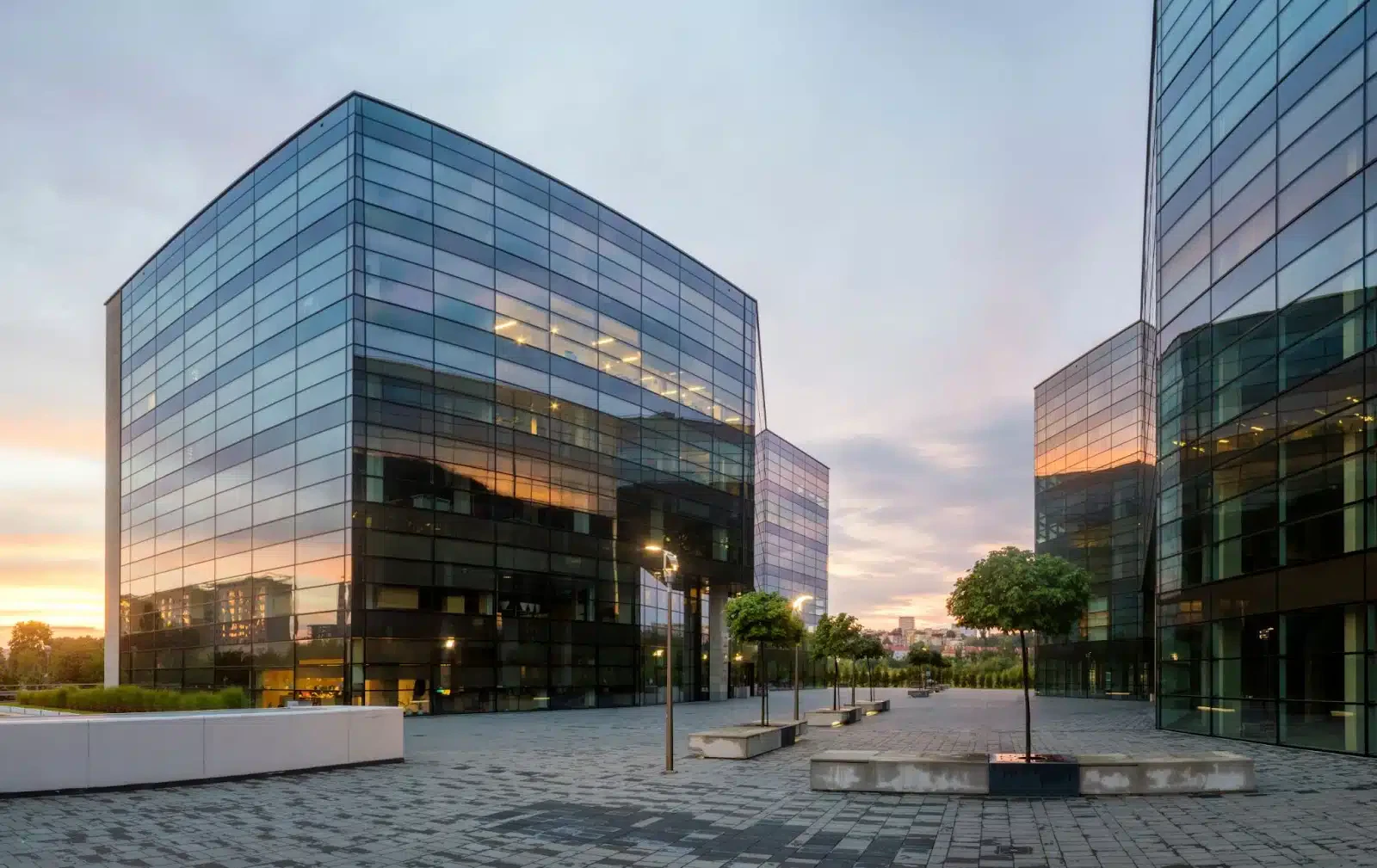 Modern glass office buildings at sunset with a paved courtyard and small trees.