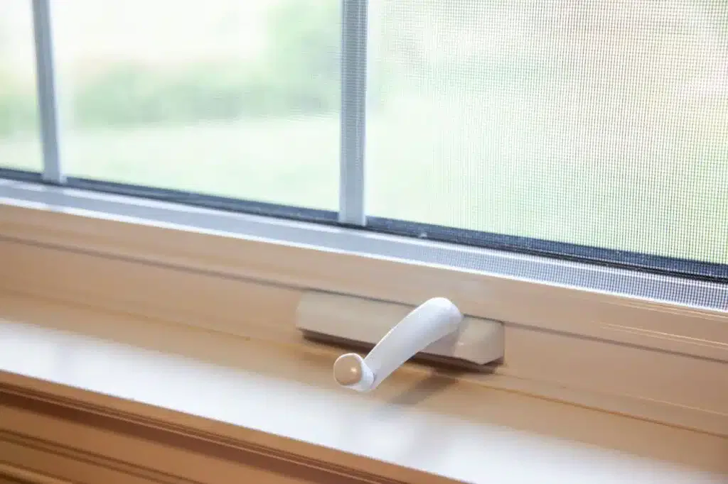 Close-up of a white window crank handle with a screen and greenery outside.